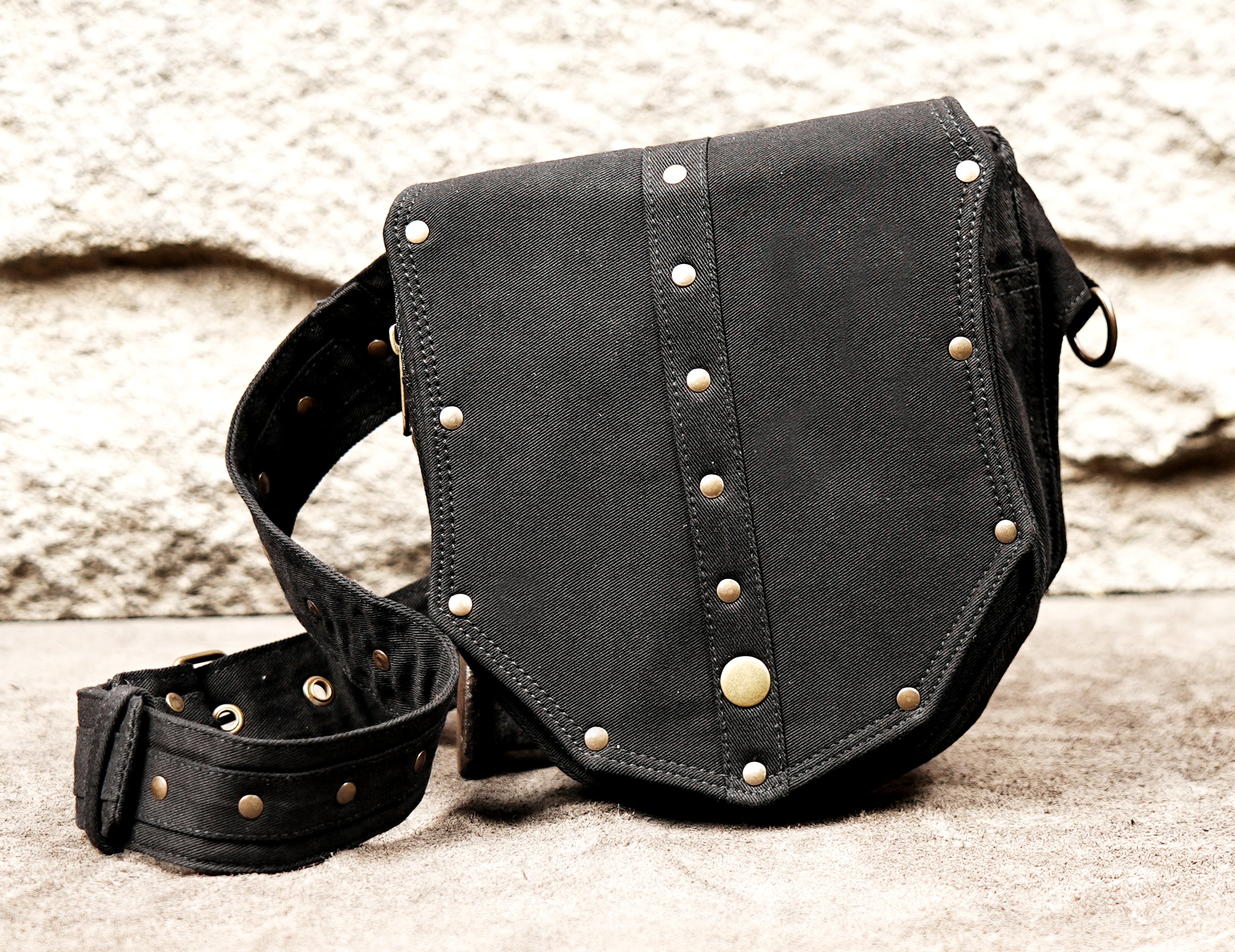Close-up view of a black cotton canvas utility belt bag showing the shield-shaped front pocket, intricate antique brass studding, and metal D-ring.