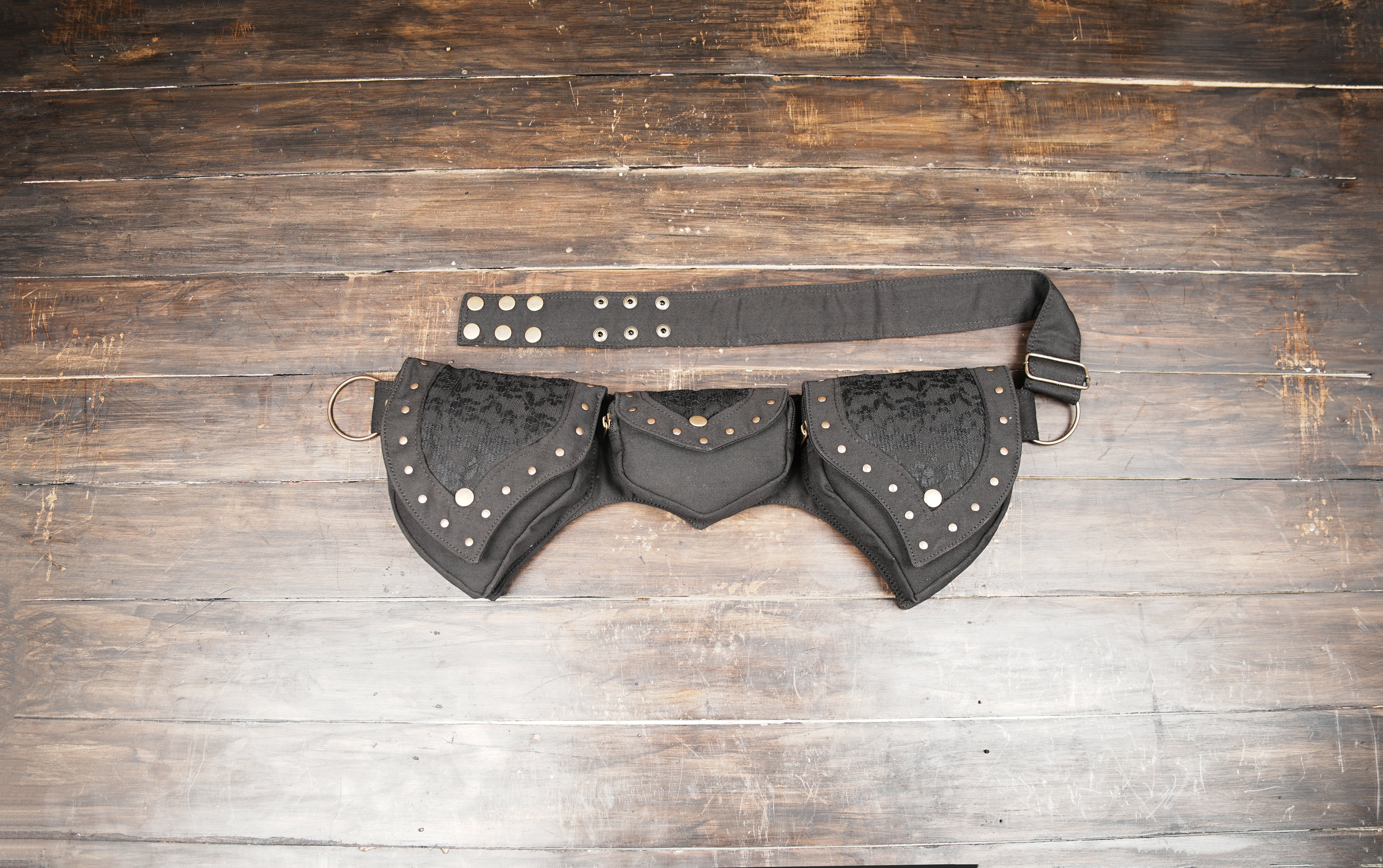 Handmade canvas pixie utility belt with dark lace trim and studded pockets laid flat on dark wood
