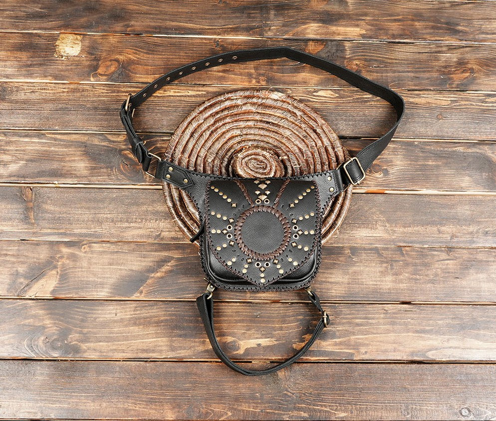 Handmade black leather leg holster bag with brass studs and circular stitching on wooden background