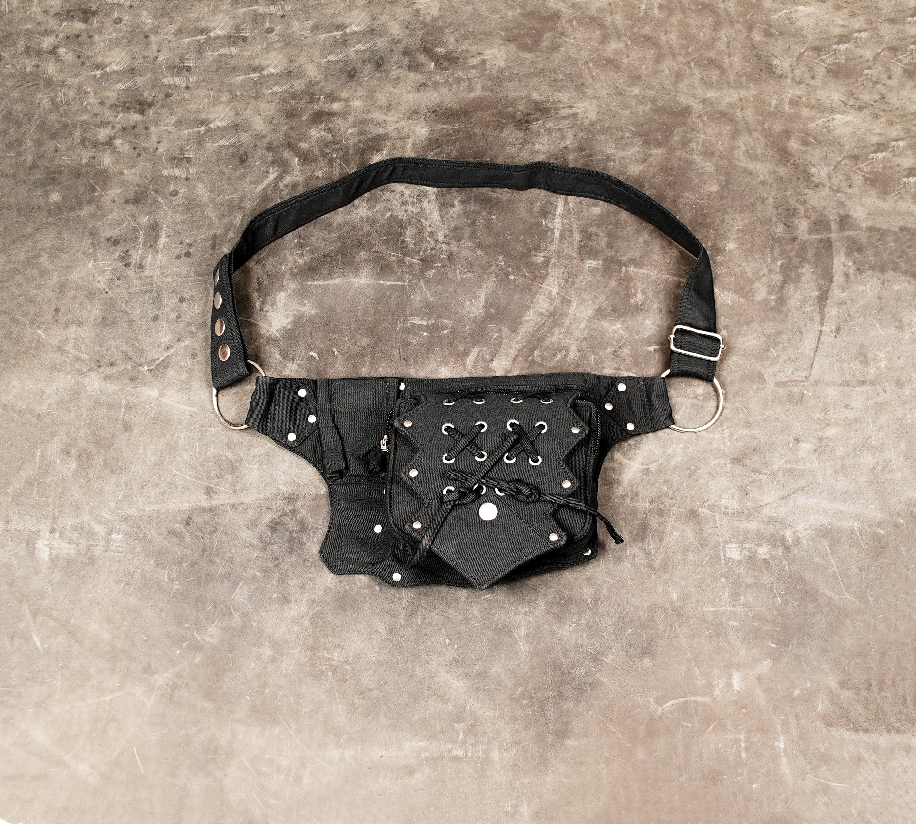 Black cotton fanny pack with unique central lace-up pocket and metal rings, laid on concrete.