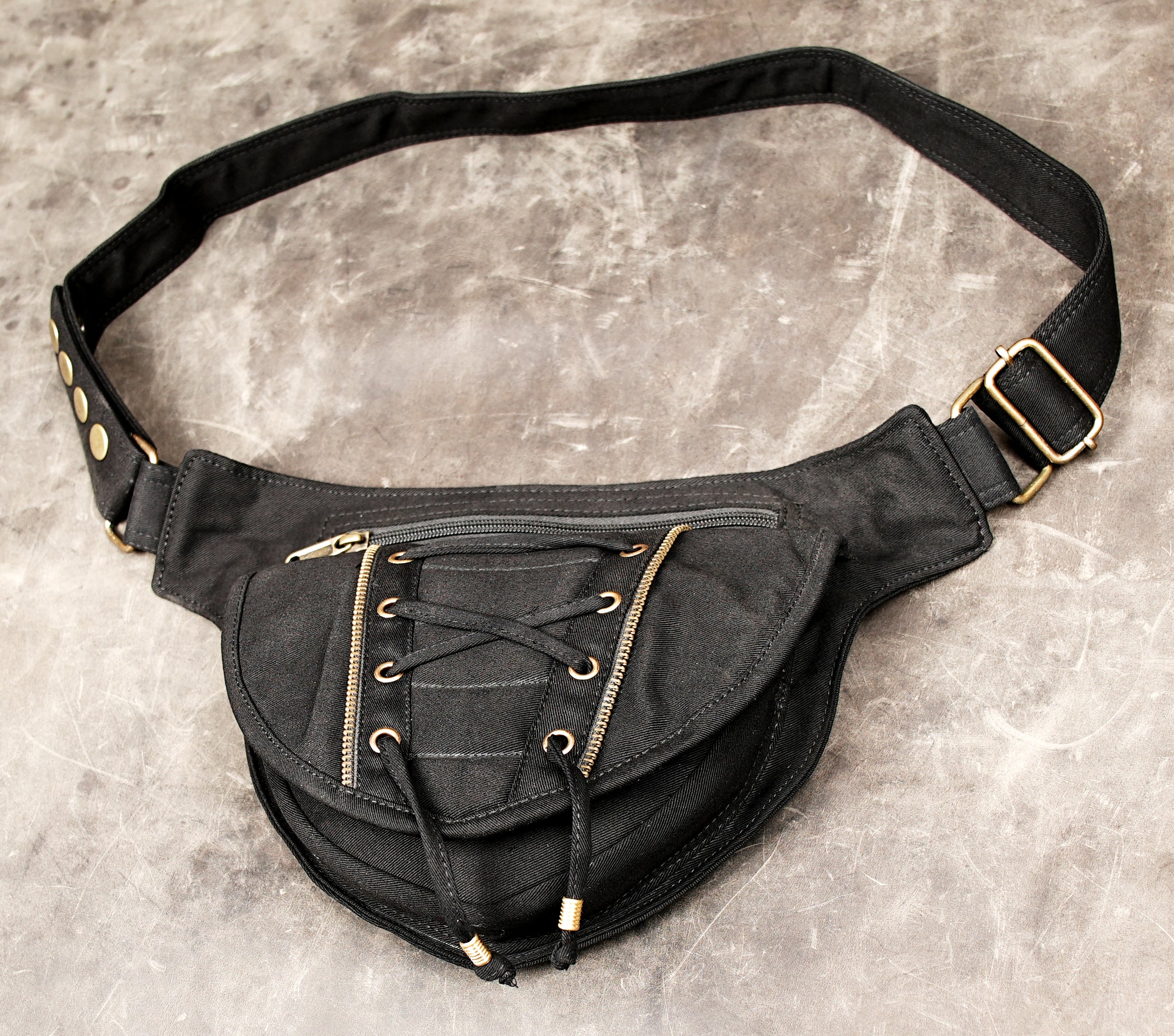 Product photo of a unisex black utility pouch with a geometric shaped flap, brass buckle details, and sturdy silver hardware, resting on a light background.