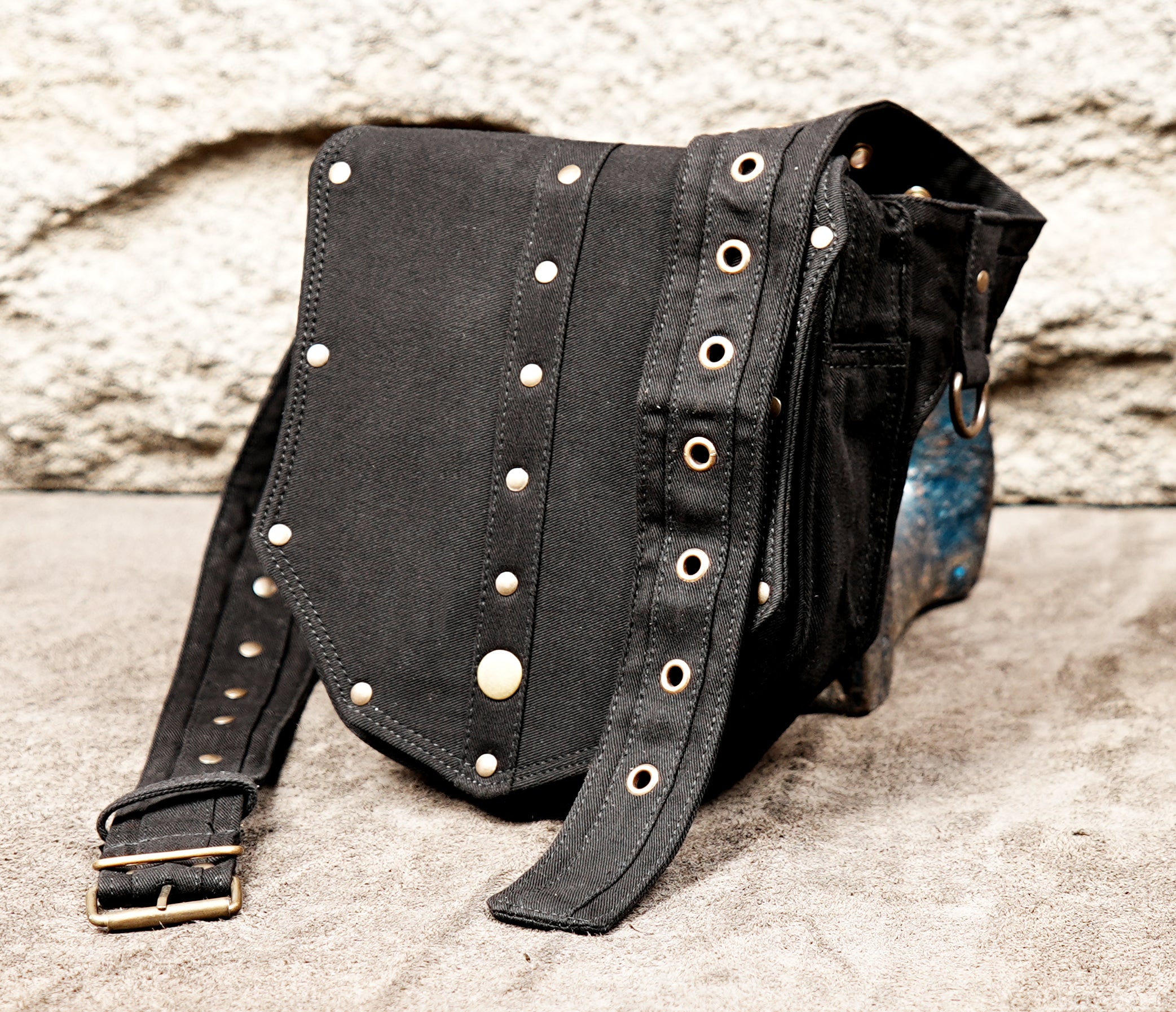 Product photo of a unisex black utility pouch with a geometric studded flap and sturdy brass buckle, resting on a wooden background.