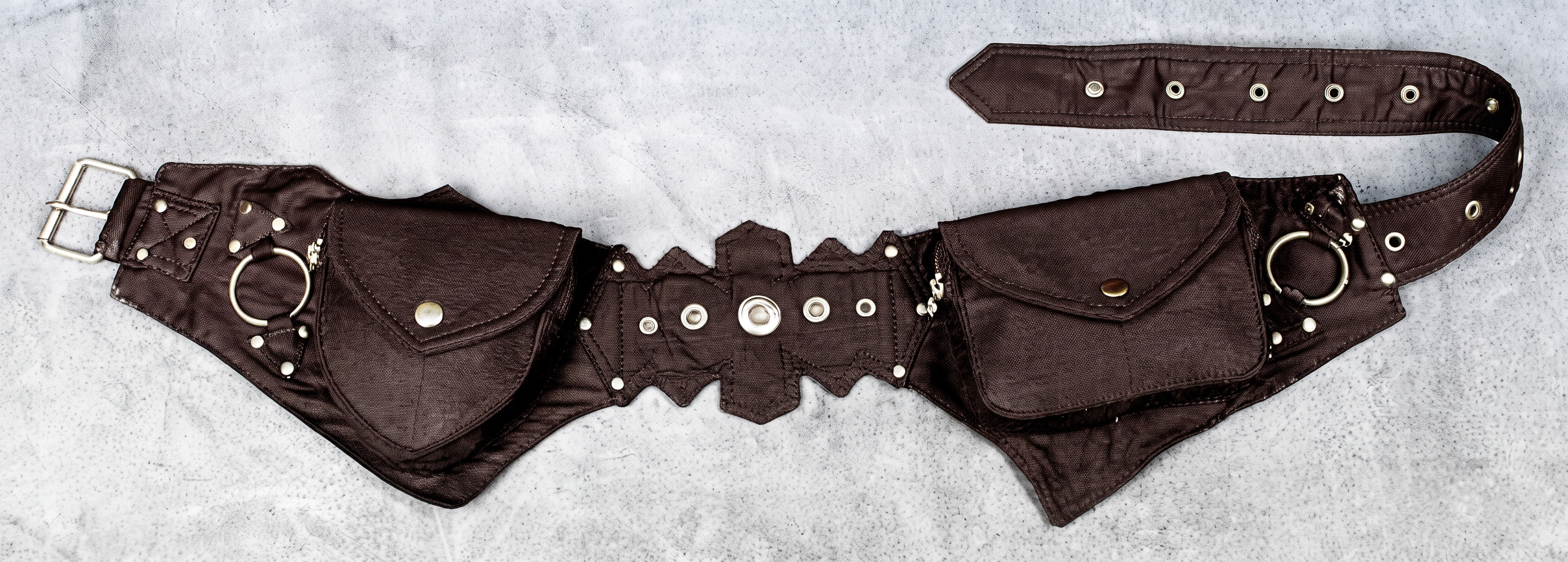 A black leather utility belt with multiple pouches, silver metal rings, studs, and a buckle, displayed flat on a textured beige background.