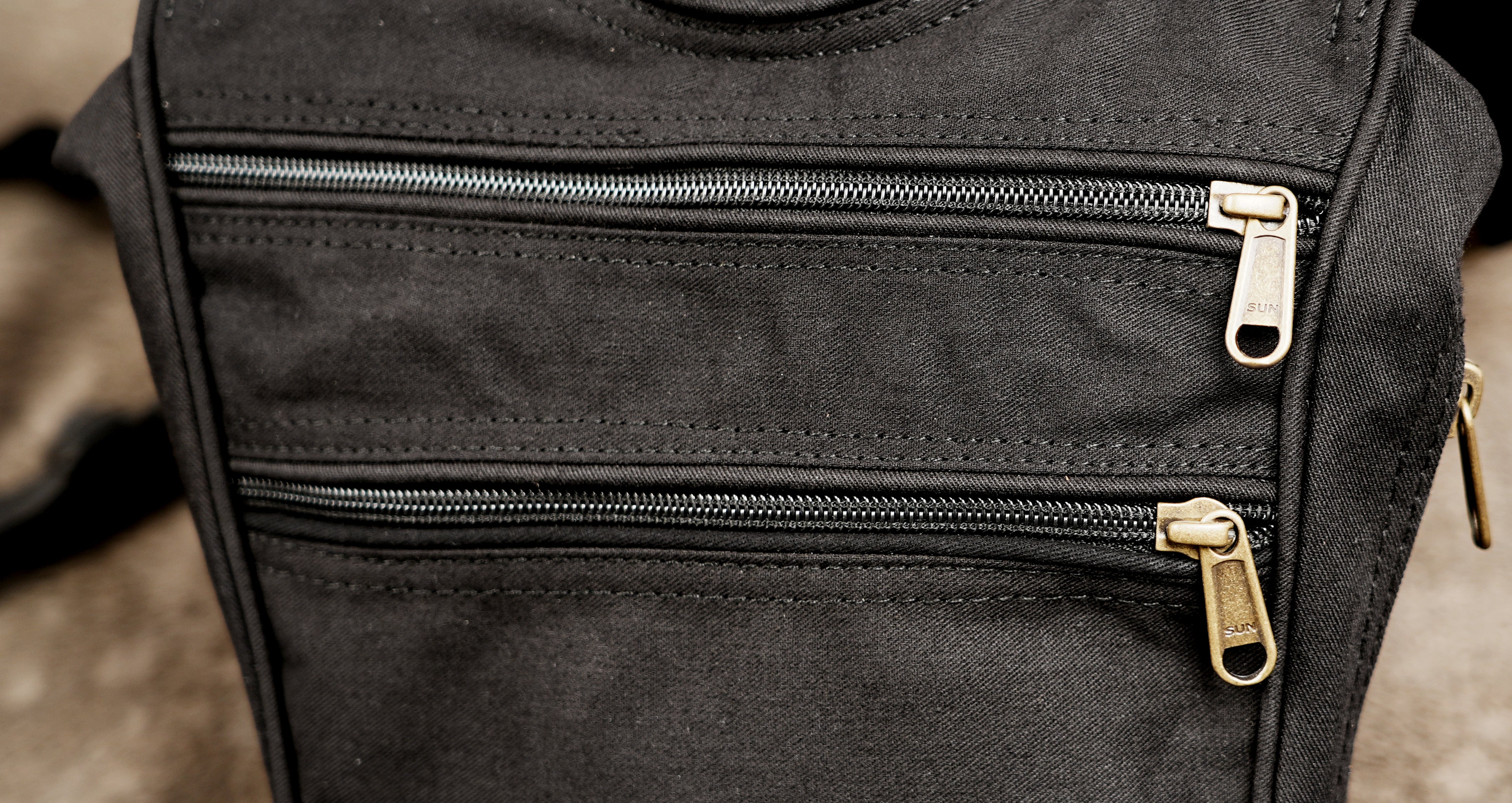 Close-up of a black canvas bag with two horizontal zippered pockets featuring gold-tone metal zipper pulls.