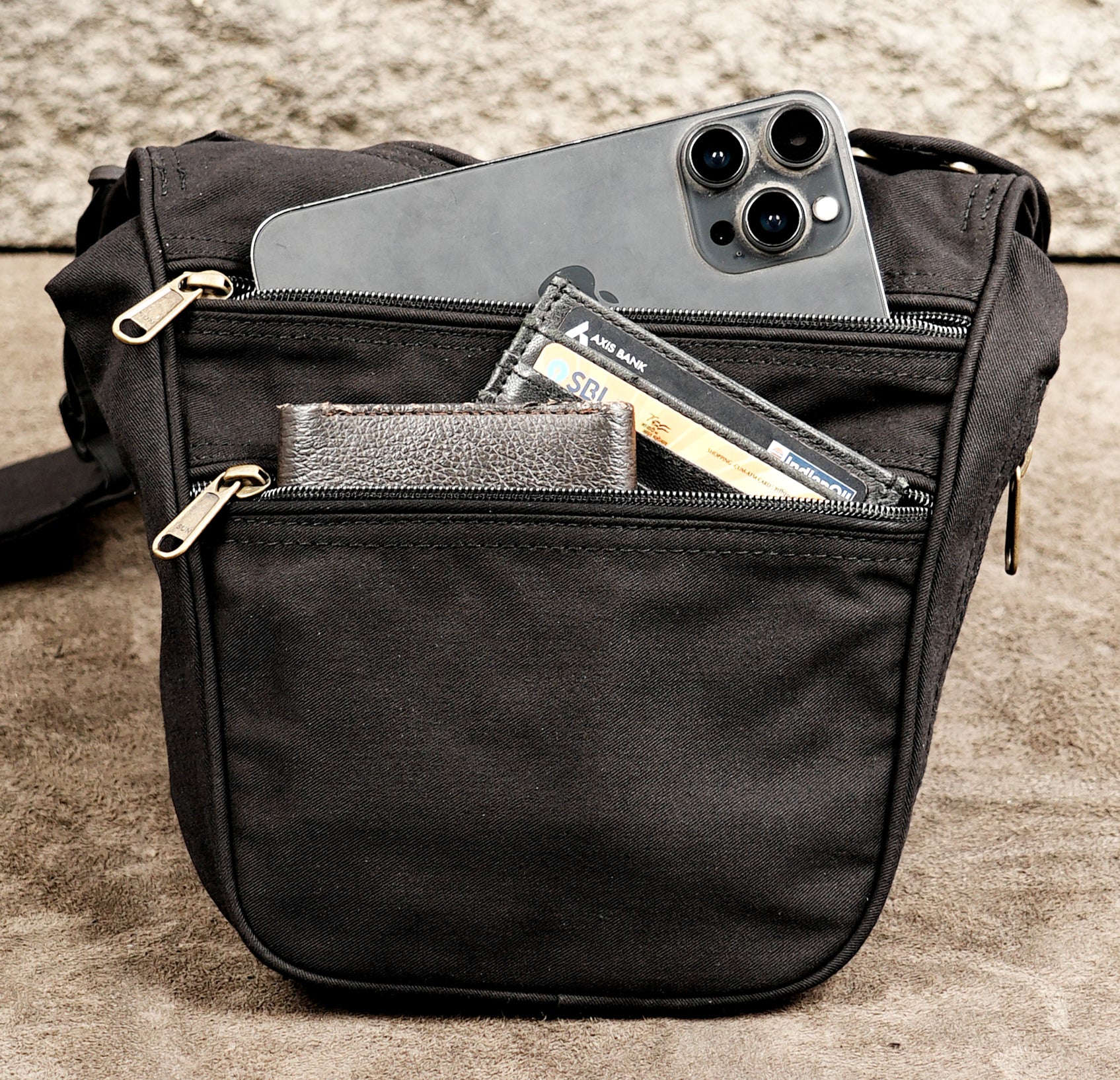 A black canvas crossbody bag with multiple zippered pockets, shown holding a smartphone, wallet, and cards.