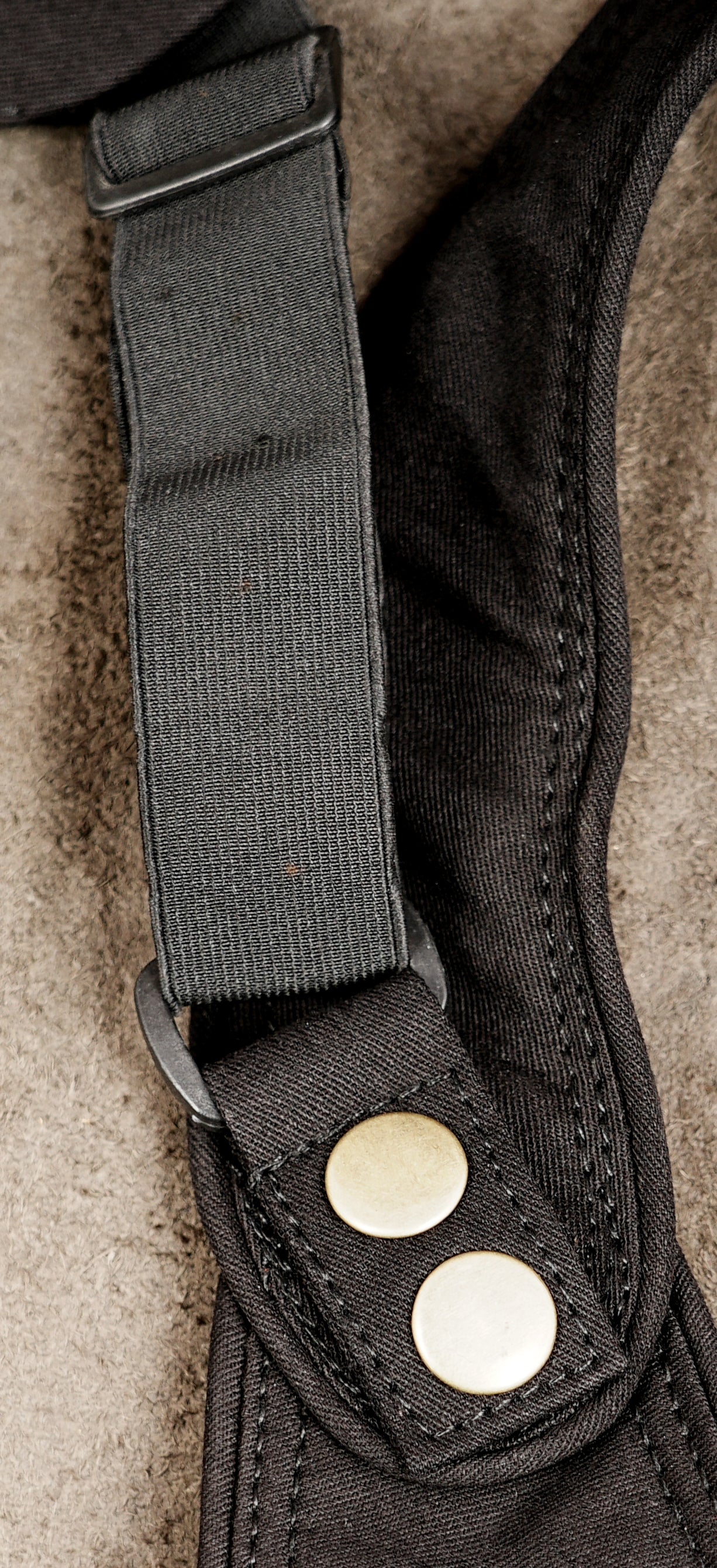 Close-up of a black fabric strap with a metal buckle and two round brass snap buttons on a textured beige surface.