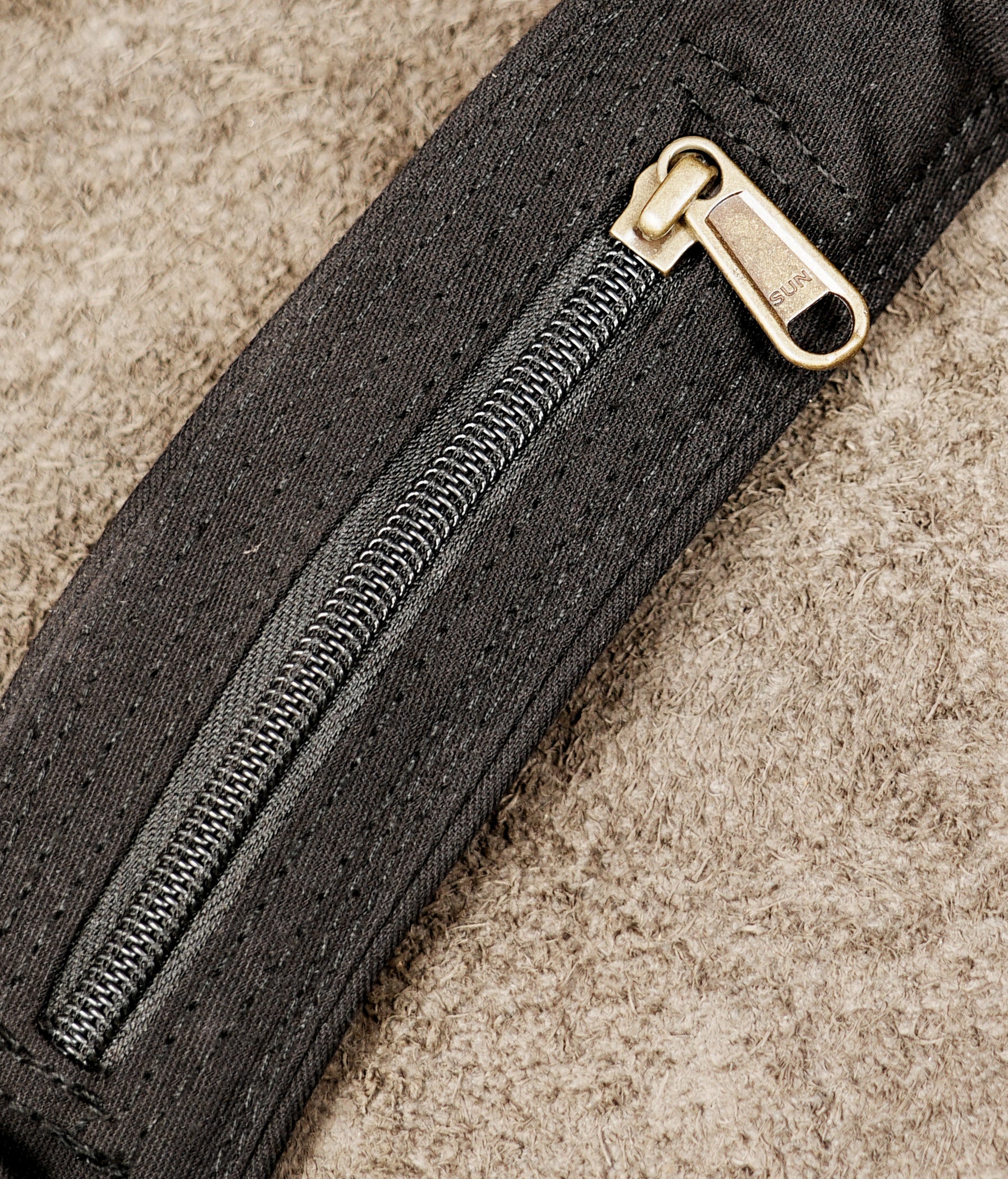 Close-up of a black textured fabric zipper with a gold-toned metal pull tab, set against a beige textured background.
