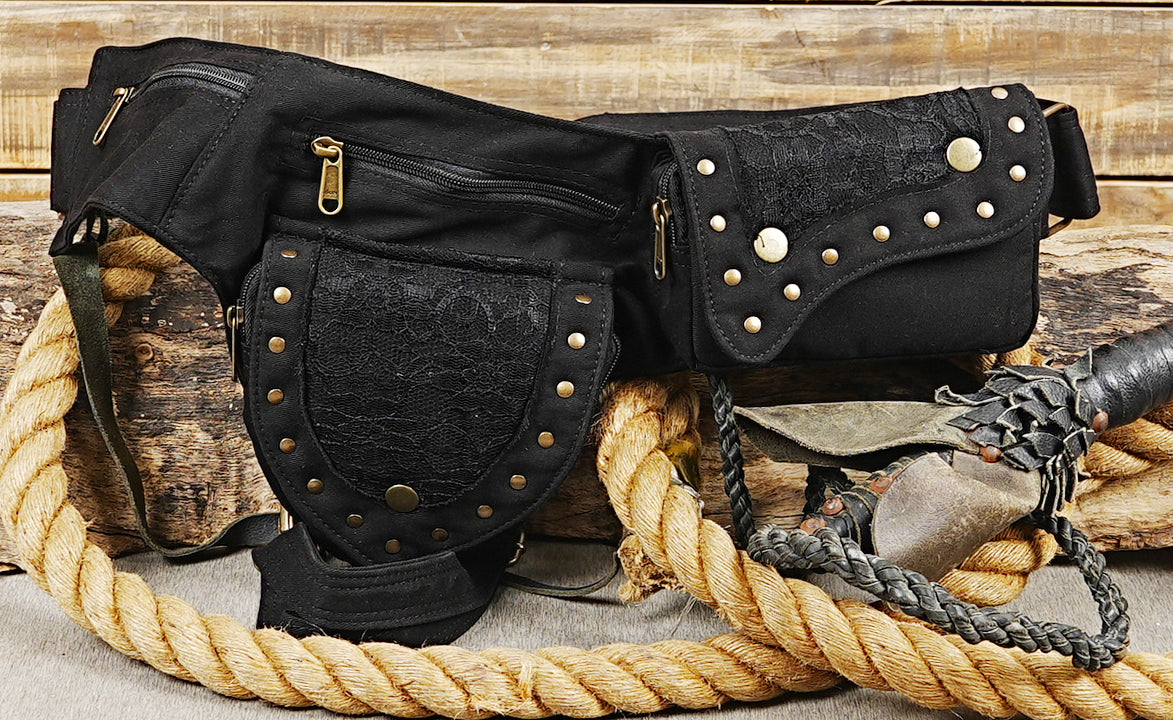 Handmade cotton canvas drop leg bag with dark lace overlays and brass studs laid on rustic wood and rope.