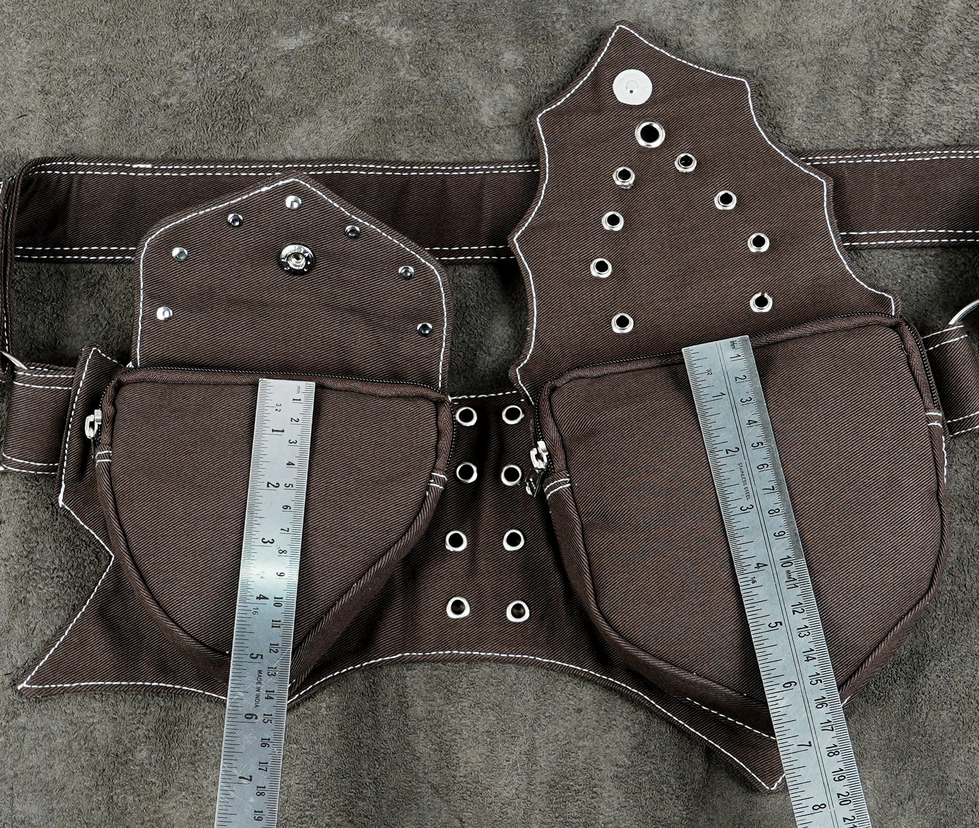 A brown fabric harness or waist belt laid flat on a textured gray surface, with metal rulers placed vertically over the front pockets to show measurements. The harness has white contrast stitching, silver metal grommets, and small silver studs.
