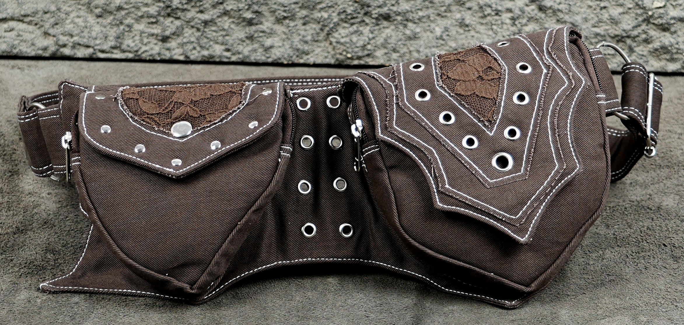 A dark brown fabric belt bag with silver metal grommets, white contrast stitching, and brown lace appliques on the front pockets.