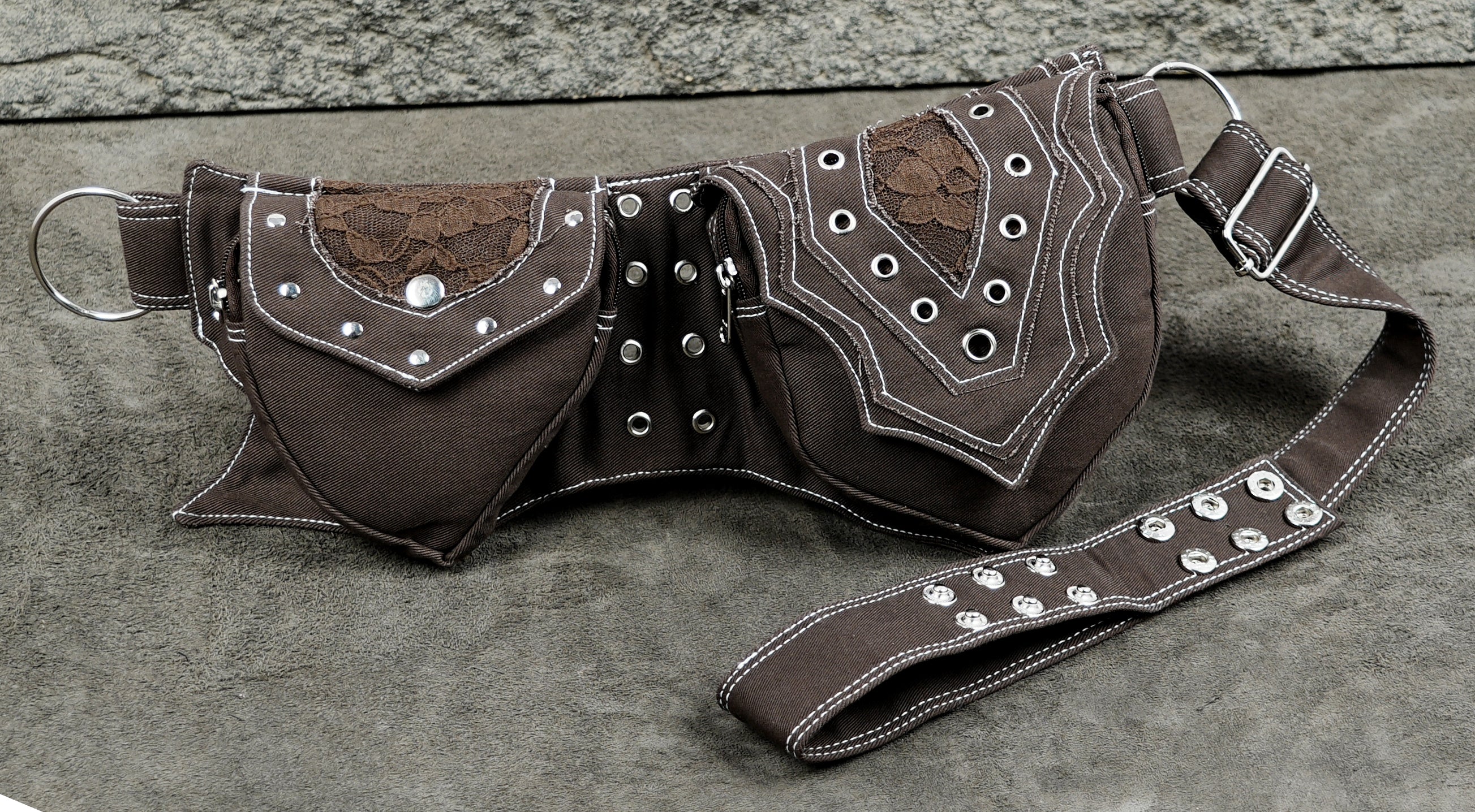 A dark brown fanny pack with western-style stitching, silver grommets, lace applique, and a matching adjustable strap, displayed on a gray textured surface.