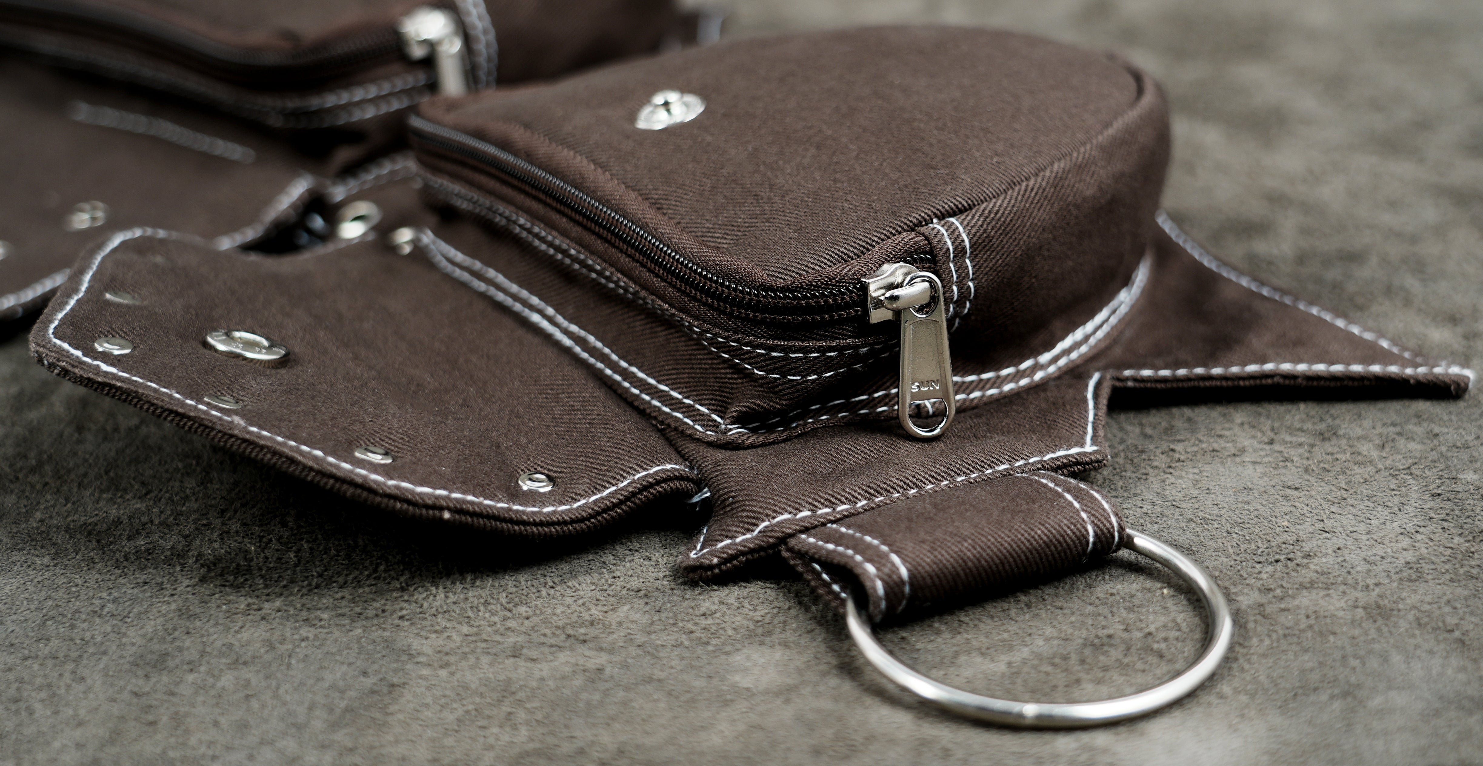 A close-up of a dark brown canvas and pouch with white contrast stitching, silver metal hardware, and a key ring, resting on a textured gray surface.
